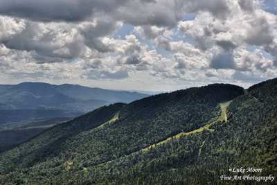 A vast, summer landscape that encompasses Stow Mountain Resort, Mount Mansfield, and Mt. Mansfield State Park in Stowe, Vermont - New England. To the left, light rays stream down through the clouds in the distance. 